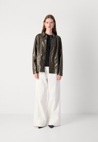 Giacca di pelle verde scuro con stampa leopardata, chiusura a bottoni e vestibilità comoda, abbinata a pantaloni bianchi larghi e scarpe nere.
