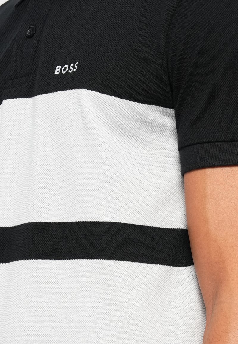 Polo a rayas en blanco y negro, hecho de tejido texturizado, con un collar y botones negros, y el logo "BOSS" bordado en el pecho.