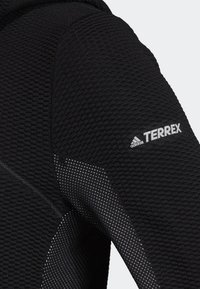 Haut à manches longues noir texturé avec motif tricoté, accents de panneaux avec petits points blancs, et logo "adidas TERREX" sur l'épaule.