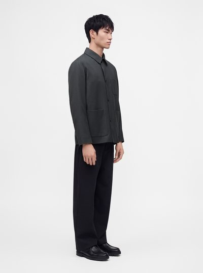 Jeune homme portant une veste sombre boutonnée avec des poches, un pantalon noir à jambes larges et des chaussures noires, debout devant un fond blanc uni.