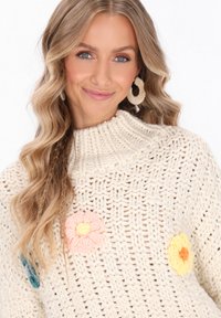 Cremfarbener gehäkelter Pullover mit hohem Kragen, der mit bunten Blumenapplikationen in Pink, Gelb und Türkis verziert ist. Strukturierter Strickmuster im gesamten Pullover.