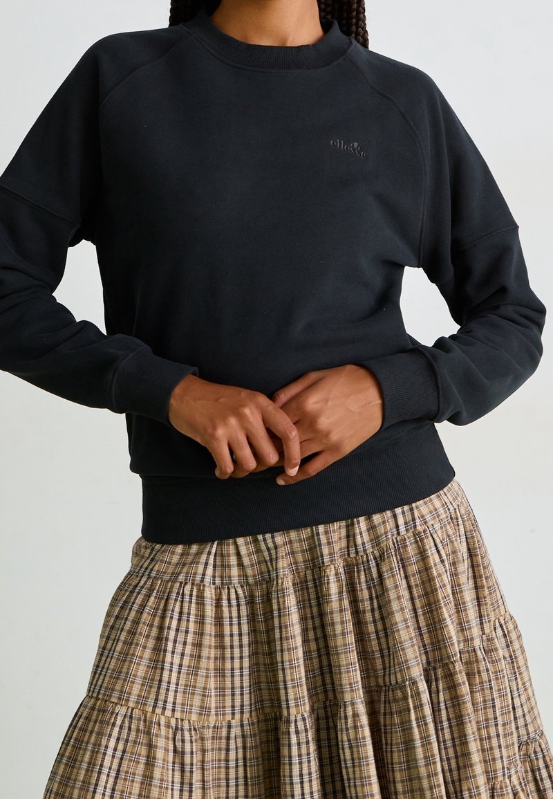 Personne portant un sweat noir et une jupe à carreaux beige, mains jointes à la taille, haut du corps visible, fond neutre.