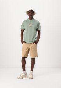 T-shirt verde de algodão com uma impressão gráfica, calções de carga bege com bolsos, ténis bege e um chapéu de balde branco. Palete de cores neutras.