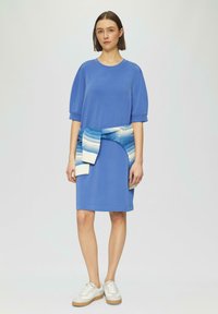 Blaues kurzärmliges Kleid mit rundem Ausschnitt und lockerer Passform, akzentuiert durch einen blau-weißen gestrickten Wickelgürtel in der Taille. Weiße Sneakers.