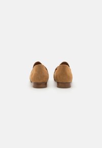 Mocasines de ante en color marrón claro con un acabado suave, tacón de madera bajo y diseño de parte trasera redondeada; presentan costuras mínimas y sin herrajes visibles.