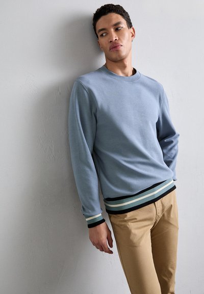 Light blue sweatshirt with long sleeves, textured fabric, and striped black, green, and white ribbed hem. Paired with beige chinos.