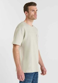 Beige t-shirt met korte mouwen, voorzien van een gestructureerde afwerking en een borstzak, gecombineerd met blauwe denim jeans, zijaanzicht.