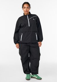 Musta tuulitakki, jossa on vetoketjullinen rintatasku ja heijastavia yksityiskohtia, yhdistettynä väljiin mustiin housuihin ja vihreisiin urheilukenkiin.