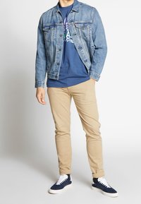 Chaqueta de mezclilla, de color azul claro, con cierres de botón y dos bolsillos en el pecho. Camisa azul marino debajo. Pantalones caqui y zapatillas azul marino con suelas blancas.