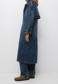 Veste en jean oversize de couleur bleue avec une coupe longue et des poches devant ; portée sur un sweat à capuche marron, associée à un jean large et des chaussures marron.