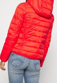 Persona che indossa una giacca a vento rossa con cappuccio e jeans aderenti blu chiaro, vista da dietro su uno sfondo semplice.