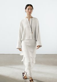 Chemise en lin gris clair avec des détails de boutons et des manches larges, associée à une jupe blanche à volants superposés ; sandales marron en guise de chaussures.