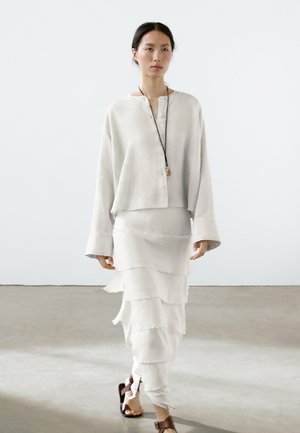 Camicia di lino grigio chiaro con dettagli a bottoni e maniche larghe, abbinata a una gonna bianca stratificata con volant; sandali marroni come calzature.