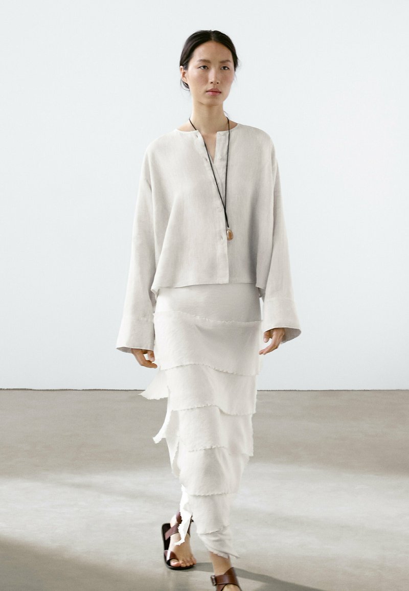 Chemise en lin gris clair avec des détails de boutons et des manches larges, associée à une jupe blanche à volants superposés ; sandales marron en guise de chaussures.