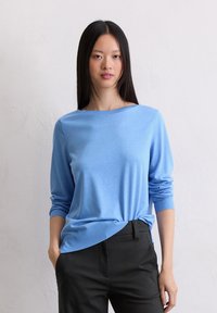 Helles blaues Langarmshirt mit Rundhalsausschnitt, das eine geknotete Detail an der Taille aufweist, kombiniert mit schwarzen Maßanzugshosen. Glatte Textur.