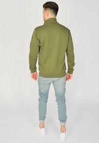 Oliwkowa polarowa kurtka z wysokim kołnierzem, prążkowanymi mankietami i dołem; zestawiona z jasnoniebieskimi slim fit dżinsami i białymi sneakersami.