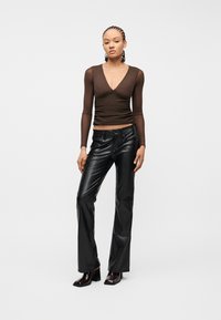 Γυναίκα με πλεξούδα, ντυμένη με στενό διάφανο καφέ μακρυμάνικο τοπ, μαύρο δερμάτινο παντελόνι και σκούρα καφέ μποτάκια με τακούνι, στέκεται μπροστά σε μονόχρωμο φόντο.