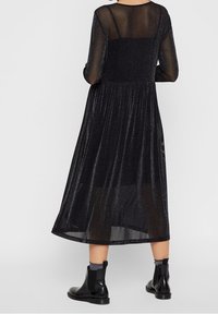 Vestido midi negro y brillante con mangas largas de tul y un corsé ajustado, que fluye hacia una falda plisada. Llevado con botines negros.