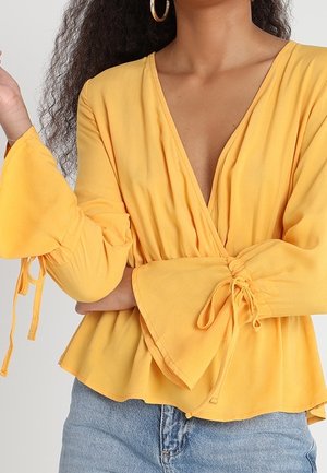 Blusa amarilla tipo envoltura con escote en V profundo, mangas acampanadas y lazos. Hecha de una tela ligera, con una textura suave y un dobladillo acampanado.