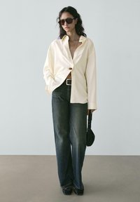 Camicia gialla chiaro con bottoni, vestibilità larga. Jeans a gamba larga blu scuro, orlo sfrangiato. Occhiali da sole neri e borsetta piccola. Texture lisce.