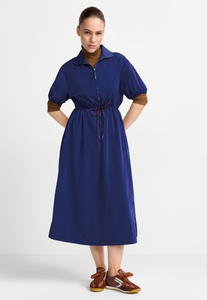 Frau trägt ein marineblaues Midikleid mit Puffärmeln, darunter ein braunes Langarmshirt, und braune Sneakers, steht mit den Händen hinter dem Rücken.