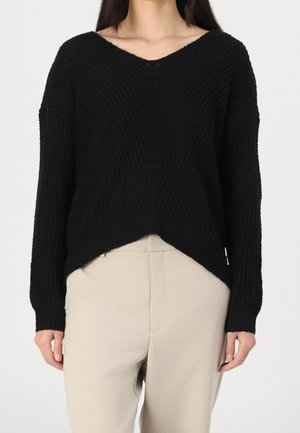 Vrouw draagt een losse, zwart geribde trui met V-hals en lange mouwen en een beige broek met hoge taille, staand tegen een effen achtergrond.
