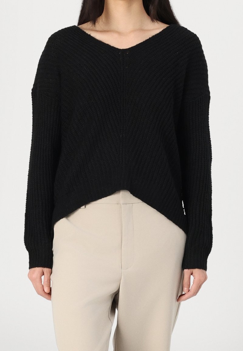 Femme portant un pull noir à côtes, ample, à col en V et manches longues, avec un pantalon beige taille haute, debout devant un fond uni.