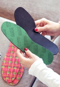 Two insoles are held in hands: one black with perforations and one green with textured carbon print. A patterned insole is visible in the background.