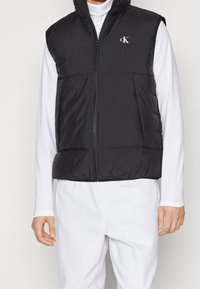 Schwarze Steppweste mit Reißverschluss vorn, geripptem Muster und Logo auf der Brust. Wird über einem weißen Langarmshirt mit hohem Kragen getragen.