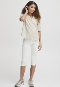 Camisa de lino color beige claro con escote en V y tres botones, combinada con leggings capri blancos y zapatillas grises, destacando un estilo casual.