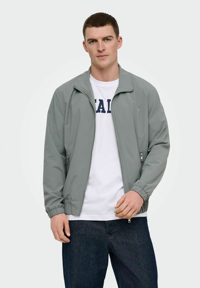 Fiatal férfi szürke cipzáros dzsekiben, fehér, szöveges pólóval és sötétkék farmernadrágban, egyszerű háttér előtt állva.