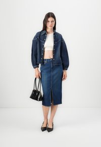Ensemble en jean comprenant une veste et une jupe de couleur bleu foncé, avec des manches bouffantes, une fente à l'avant et une silhouette ajustée. Accompagné d'un sac à main noir.