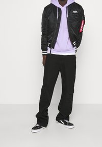 Černá bomber bunda s bílými pruhy, lilková mikina, černé cargo kalhoty a černé tenisky Vans. Tkanina se zdá být hladká a odolná.