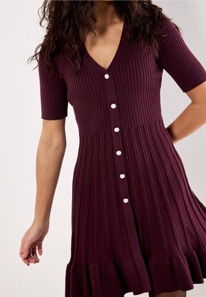 Vrouw draagt een geribbelde, korte mouw, geplooide pruimkleurige jurk met knopen en een uitlopende zoom, staand tegen een effen lichte achtergrond.