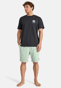 T-shirt en coton noir avec un motif floral sur la poitrine gauche, associé à un short texturé vert clair. Pieds nus, debout devant un fond blanc.
