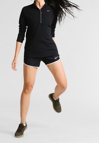 Nainen hyppää mustassa Nike pitkähihasessa paidassa, mustissa Nike lyhyissä housuissa valkoisilla yksityiskohdilla ja oliivinvihreissä urheilukengissä.