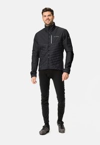 Zwarte geïsoleerde jas met een gewatteerde voorkant, hoge kraag, ritssluiting aan de voorkant en een nauwsluitende pasvorm. Draag het over een strakke zwarte broek en fietschoenen.