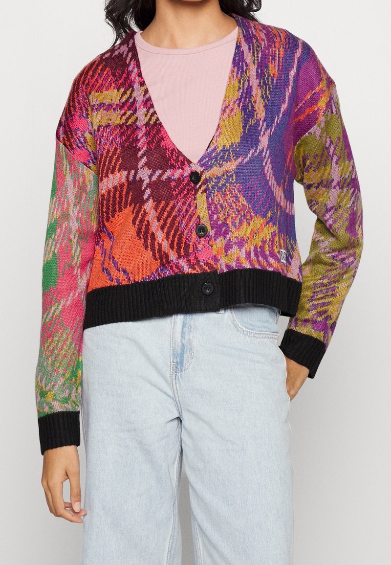Kobieta ubrana w kardigan w wielokolorowy abstrakcyjny wzór z czarnymi guzikami i wykończeniem, na jasnoróżowej koszuli i jasnoniebieskich dżinsach.