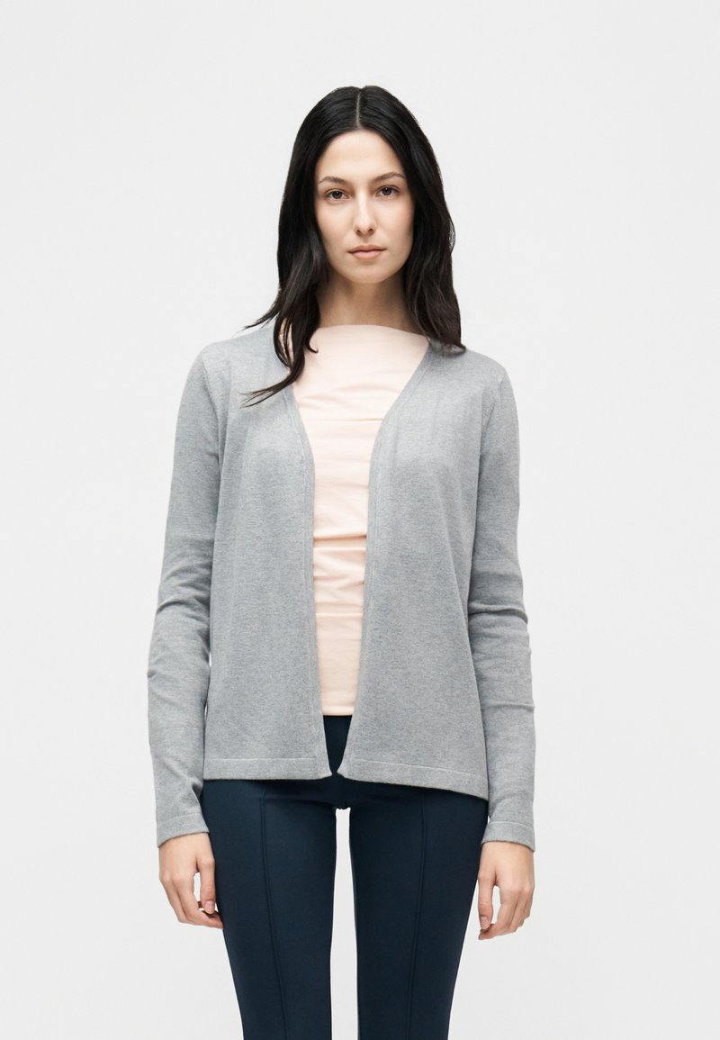 Γυναίκα με μακριά σκουρόχρωμα μαλλιά φοράει ροζ ανοιχτό τοπ, γκρι ζακέτα ανοιχτού μπροστινού μέρους και σκουρό μπλε στενά παντελόνια, στέκεται μπροστά σε απλό φόντο.