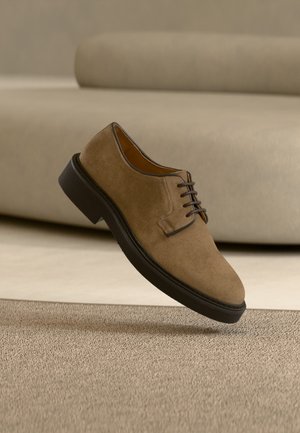 Καφέ δερμάτινο παπούτσι φόρμας με κορδόνια και μαύρη σόλα να αιωρείται πάνω από μπεζ υφή χαλιού και φόντο από καναπέ με το ίδιο χρώμα.