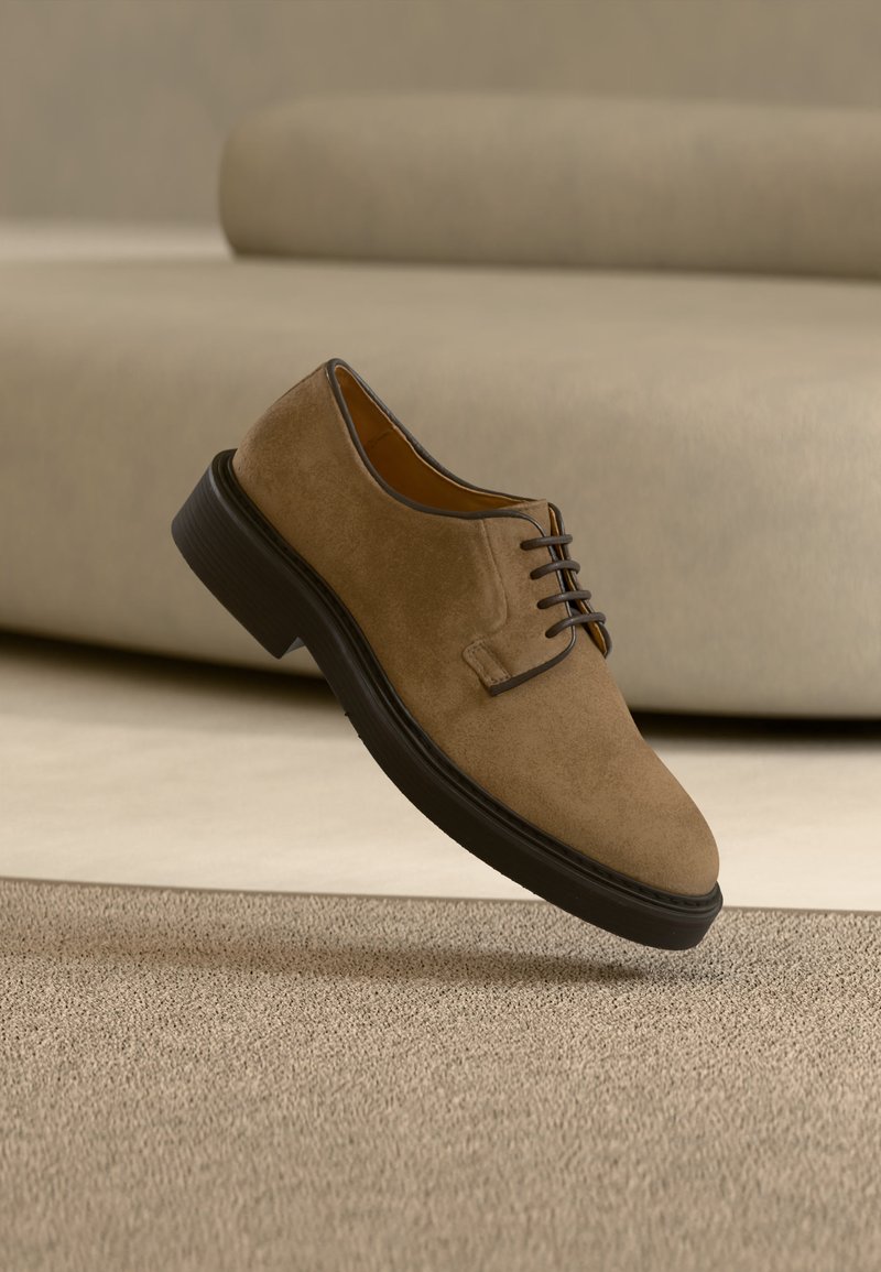 Καφέ δερμάτινο παπούτσι φόρμας με κορδόνια και μαύρη σόλα να αιωρείται πάνω από μπεζ υφή χαλιού και φόντο από καναπέ με το ίδιο χρώμα.