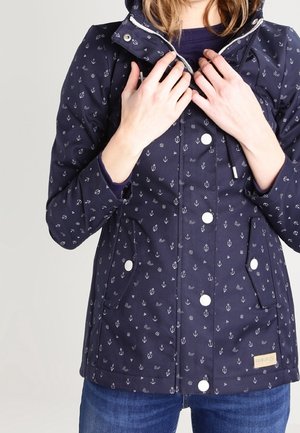 Vrouw draagt een marineblauwe jas met kleine witte ankerpatronen, een knoopsluiting aan de voorkant en klepzakken, ze houdt de kraag bij de rits vast.