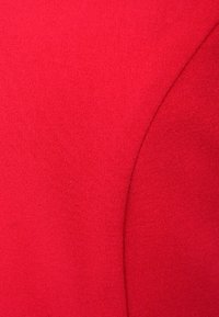 Czerwona tkanina o gładkiej fakturze i subtelnym połysku, z wydatnym detalem zakrzywionego szwu. Materiał wydaje się być trwały i mocno tkany.