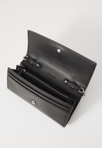 Czarny portfel ze skóry o gładkiej teksturze, wyposażony w komorę na zamek, kilka przegródek na karty oraz zapięcie na zatrzask. Wewnątrz widoczne logo marki.