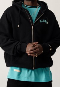 Sort zip-up hættetrøje med et teal logo, båret over en blå mesh skjorte. Bemærkelsesværdige detaljer inkluderer snore og et lille logo på skjorten.