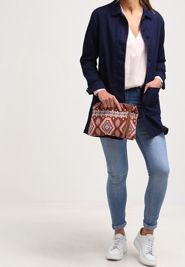 Giacca camicia blu navy, blusa rosa chiaro, jeans blu e sneakers bianchi. Tiene una pochette rossa con un motivo geometrico e chiusura con cerniera.