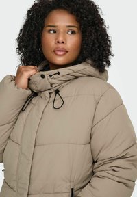 Bēša pūkaina jaka ar horizontālām šuvēm, augstu apkakli un regulējamām auklām. Ir rāvējslēdzēja sānu kabatas un gluda tekstūra.