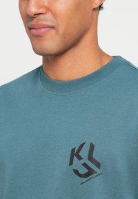 Tirkizni dukserica s okruglim izrezom od mekog materijala s crnim logotipom na lijevoj strani prsa. Glatka tekstura i minimalistički stil.