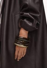 Dunkelbrauner Leder mantel mit gerafften Ärmeln. Die Hand geschmückt mit drei klobigen Armbändern: zwei schwarze und eines metallisch goldfarben, mit glatter Textur.