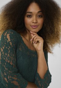 Green lace top with three-quarter sleeves, featuring floral patterns and a scalloped neckline. The texture appears delicate and intricate.